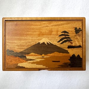May include: A rectangular wooden box featuring an intricate marquetry inlay on the lid. The design depicts a landscape with a snow-capped mountain, pine trees, and small boats on water, rendered in various shades of natural wood grain and tones.