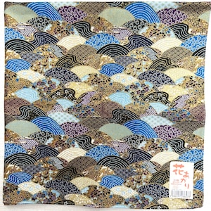 Op de afbeelding: Vierkante stof met een herhalend patroon van gestileerde golven in blauw, goud, zwart, wit en paars. Het ontwerp doet denken aan traditionele Japanse kunst. Een klein wit label met Japanse tekst staat in de rechter benedenhoek.