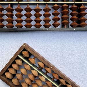 Puede incluir: Dos ábacos de madera con cuentas y marcos de color marrón oscuro. Un ábaco tiene un marco gris oscuro y el otro un marco marrón. Las cuentas tienen forma de cono y están dispuestas en filas. Los ábacos están sobre un fondo blanco.