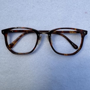 Puede incluir: Gafas con montura de carey. Las gafas tienen forma redondeada y puente metálico. Las lentes son transparentes. La montura es de color marrón oscuro y ámbar.