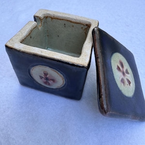 May include: A square ceramic box with a lid, in a dark blue color. The lid is off to the side. The box and lid have a floral design in the center. The interior of the box is a light green color.