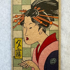 以下が含まれることがあります： 伝統的な衣装を着た女性のヴィンテージ日本の木版画。このアートワークは、オレンジ色のヘアピンで飾られた黒髪の女性が、ピンクと赤の着物を着ています。背景は緑とタンのチェック柄で、黄色のボックスに日本語のテキストがあります。