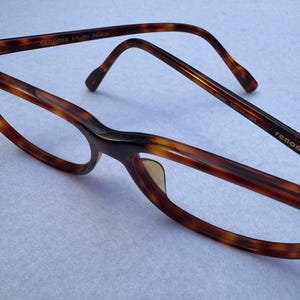 May include: Brown tortoiseshell eyeglasses with the brand name "renoma RALPH PARIS" printed on the frame. The glasses have a classic design with a rounded shape and a keyhole bridge. The frame is made of a glossy material.
