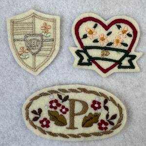 以下が含まれることがあります： 3つの刺繍パッチ。1つは盾型、もう1つは花柄のハート型、最後は文字「P」と花のディテールが入った楕円形です。すべてのパッチはクリーム色で、さまざまな色の刺繍が施されています。