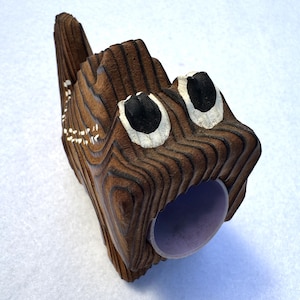 May include: A whimsical, carved wooden sculpture resembling a stylized animal head. The sculpture features a brown, textured surface with visible wood grain, black and white eyes, and a purple opening. The overall design is unique and artistic.