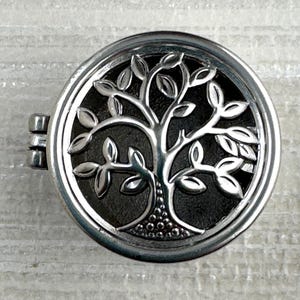 Puede incluir: Un colgante redondo plateado con cierre de bisagra. El colgante presenta un diseño detallado de árbol de la vida dentro de un marco circular. Las ramas y hojas del árbol están intrincadamente representadas sobre un fondo oscuro.