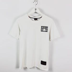 May include: White t-shirt with a crew neck and short sleeves. The shirt features a black and white logo on the chest that says "EVISU". A smaller black label with the brand name is near the bottom hem. The shirt is hanging on a white hanger.