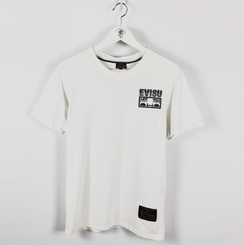 May include: White t-shirt with a crew neck and short sleeves. The shirt features a black and white logo on the chest that says "EVISU". A smaller black label with the brand name is near the bottom hem. The shirt is hanging on a white hanger.