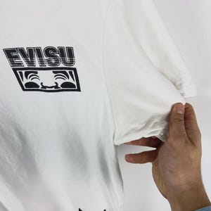 May include: White t-shirt with a black graphic print. The graphic features the word "EVISU" in a bold, stylized font above a smiling face design. The shirt is made of a soft material.