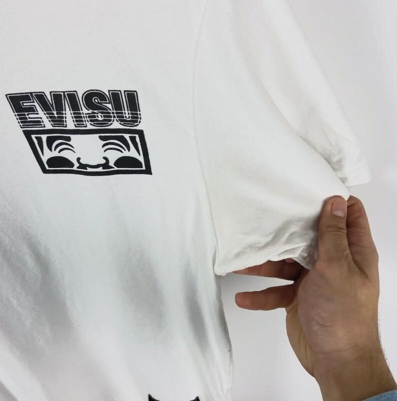 May include: White t-shirt with a black graphic print. The graphic features the word "EVISU" in a bold, stylized font above a smiling face design. The shirt is made of a soft material.