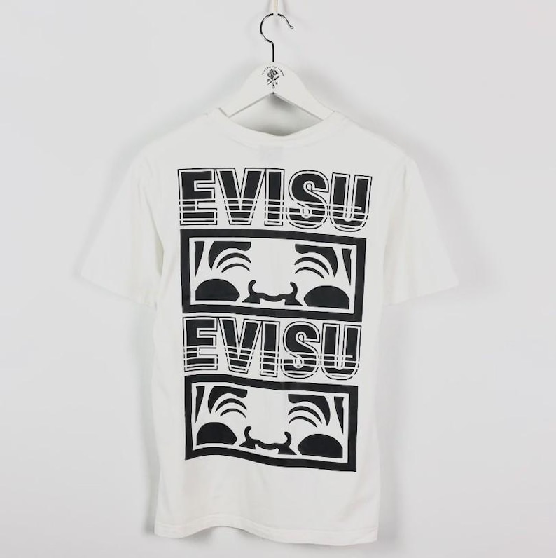 May include: White t-shirt with a black graphic design. The design features the word "EVISU" stacked twice, with a stylized face graphic in between. The shirt has short sleeves and a crew neck.