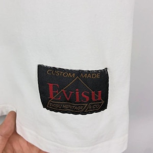 May include: Close-up of a white garment with a black label. The label reads "CUSTOM MADE" above the brand name "Evisu" in red, with "EVISU HERITAGE & CO" below. The garment's fabric is smooth.