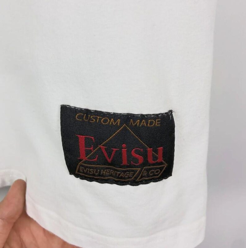 May include: Close-up of a white garment with a black label. The label reads "CUSTOM MADE" above the brand name "Evisu" in red, with "EVISU HERITAGE & CO" below. The garment's fabric is smooth.