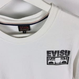 May include: White t-shirt with a crew neck and short sleeves. The shirt features the brand name "EVISU" in a black, stylized font, along with a graphic of a smiling face. The shirt has a tag with the brand name and size "M".
