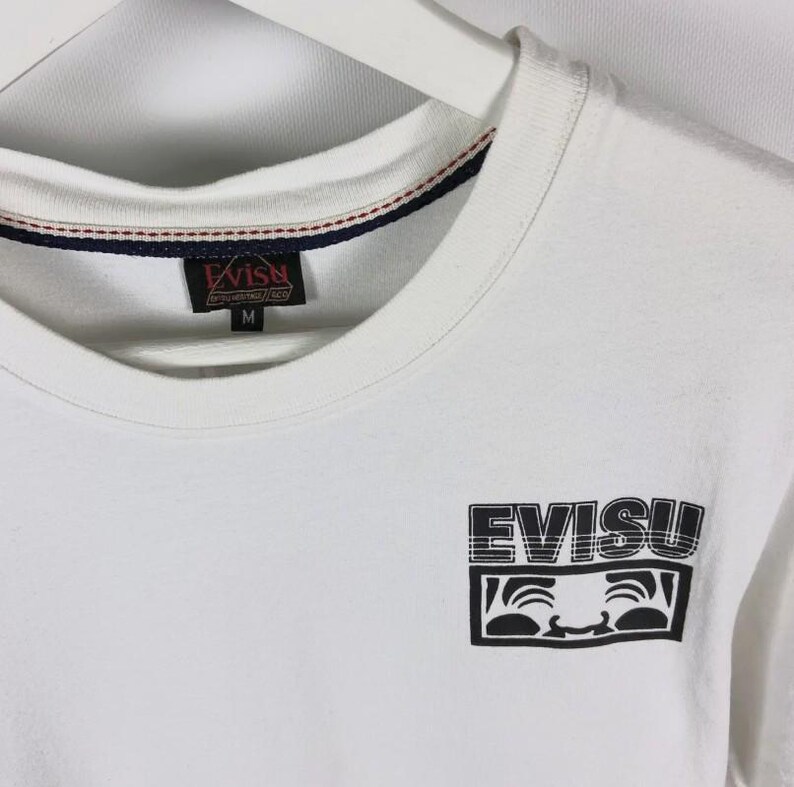 May include: White t-shirt with a crew neck and short sleeves. The shirt features the brand name "EVISU" in a black, stylized font, along with a graphic of a smiling face. The shirt has a tag with the brand name and size "M".
