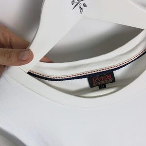 May include: A white crew neck sweatshirt with a red, white, and blue striped trim on the collar. The tag reads "Evisu" in red and gold lettering. The sweatshirt is held by a white hanger.