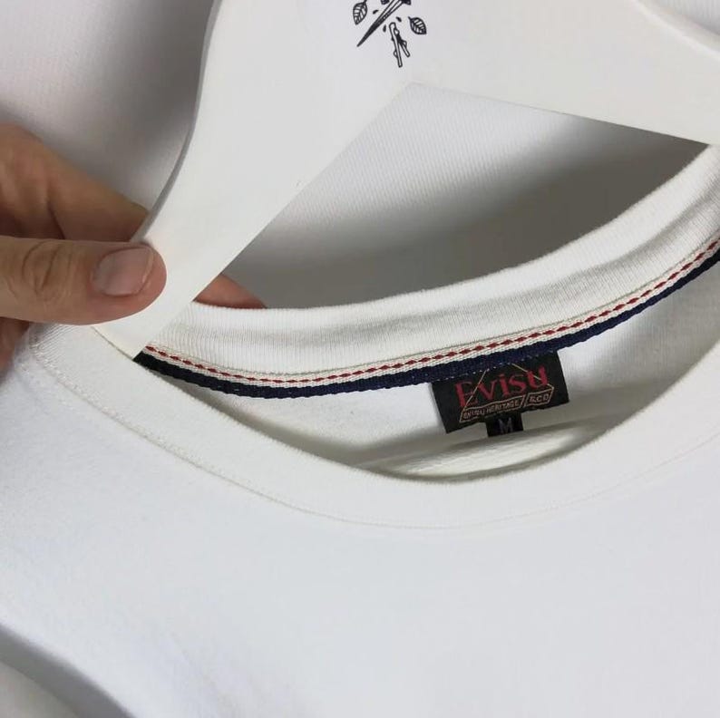 May include: A white crew neck sweatshirt with a red, white, and blue striped trim on the collar. The tag reads "Evisu" in red and gold lettering. The sweatshirt is held by a white hanger.