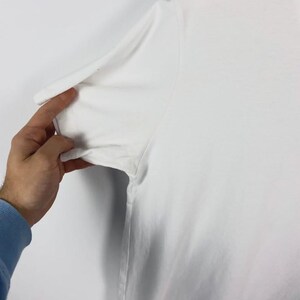 May include: Close-up of a white t-shirt sleeve. The sleeve is being held by a hand with a blue long-sleeve shirt underneath. The t-shirt appears to be made of a soft, lightweight material.