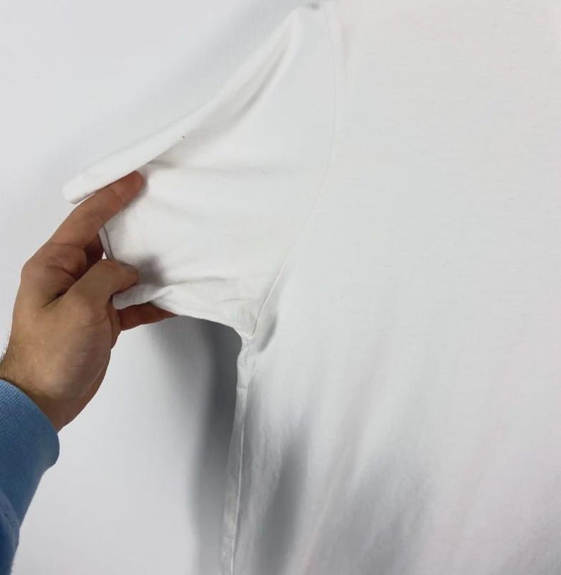 May include: Close-up of a white t-shirt sleeve. The sleeve is being held by a hand with a blue long-sleeve shirt underneath. The t-shirt appears to be made of a soft, lightweight material.