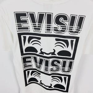May include: White t-shirt with a black graphic design. The design features the word "EVISU" stacked above a smiling face graphic, repeated twice. The shirt has a crew neck and short sleeves.