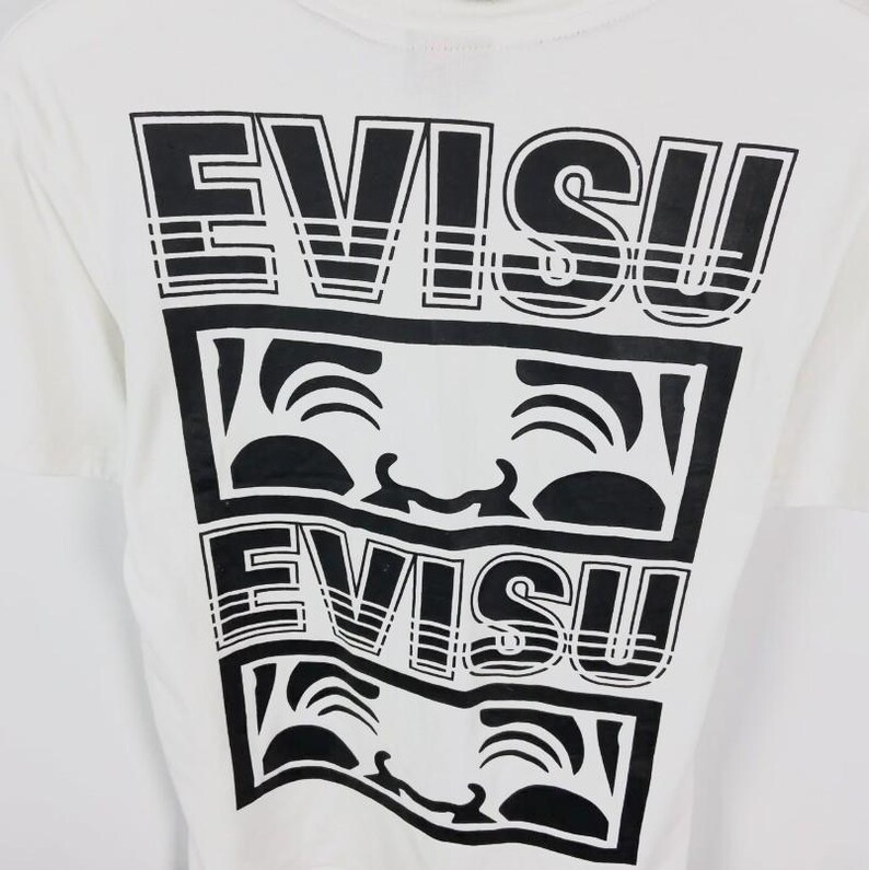 May include: White t-shirt with a black graphic design. The design features the word "EVISU" stacked above a smiling face graphic, repeated twice. The shirt has a crew neck and short sleeves.
