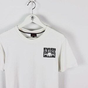 May include: White t-shirt with a crew neck and short sleeves. The shirt features a black and white graphic of a face and the word "EVISU" printed on the front. The shirt is hanging on a white hanger.