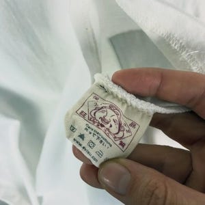 May include: Close-up of a white fabric tag with Japanese text and a cartoon face illustration. The tag includes care instructions and the website address www.evisu.com. The tag is attached to a white garment.