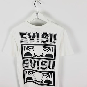 May include: White t-shirt with a black graphic design. The design features the word "EVISU" stacked above a stylized face, repeated twice. The shirt is hanging on a white hanger.