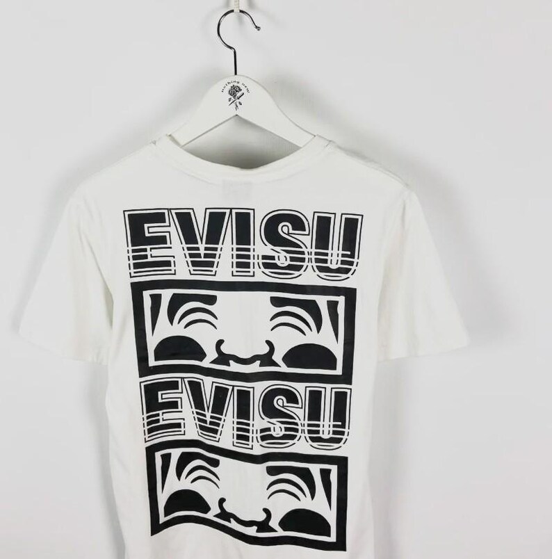 May include: White t-shirt with a black graphic design. The design features the word "EVISU" stacked above a stylized face, repeated twice. The shirt is hanging on a white hanger.