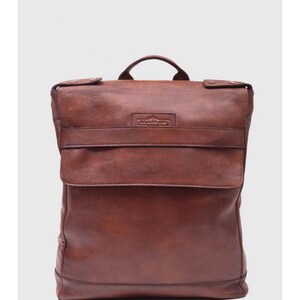 May include: A brown leather backpack with a flap closure and a top handle. The backpack has a rectangular shape with a front pocket and a small logo on the front. The leather has a distressed look.