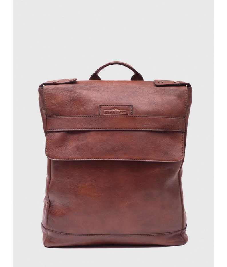 May include: A brown leather backpack with a flap closure and a top handle. The backpack has a rectangular shape with a front pocket and a small logo on the front. The leather has a distressed look.