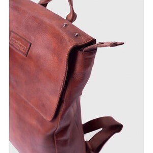 May include: A brown leather backpack with a top handle and adjustable straps. The bag features a flap closure with a buckle and metal rivets. The leather has a rich, textured appearance, suggesting durability and quality.