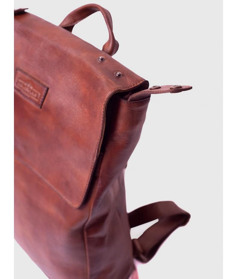May include: A brown leather backpack with a top handle and adjustable straps. The bag features a flap closure with a buckle and metal rivets. The leather has a rich, textured appearance, suggesting durability and quality.