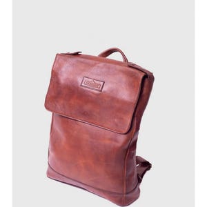 May include: A brown leather backpack with a flap closure and a top handle. The bag has adjustable straps and a small rectangular logo on the front. The leather has a distressed appearance.