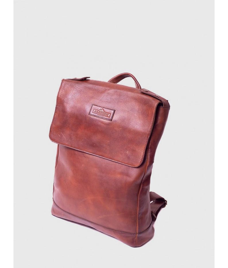 May include: A brown leather backpack with a flap closure and a top handle. The bag has adjustable straps and a small rectangular logo on the front. The leather has a distressed appearance.