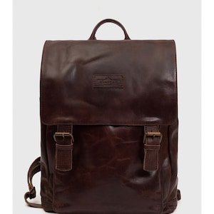 May include: A brown leather backpack with a flap closure and two buckle straps. The backpack has a top handle and adjustable shoulder straps. The brand name is embossed on a small rectangular patch on the front.