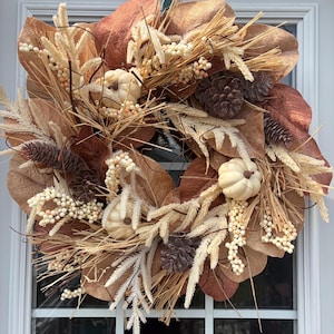 May include: A circular wreath featuring large brown magnolia leaves, cream-colored wheat stalks, small white berries, and several dark brown pinecones. Three small, off-white decorative pumpkins are nestled among the foliage and natural elements.