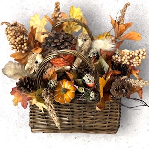 May include: A woven basket overflowing with autumn-themed decorations. The basket contains faux pumpkins, pinecones, and fall leaves in shades of orange, brown, and yellow. Decorative elements include small, furry animal figures and berry clusters.