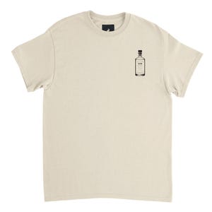 Op de afbeelding: Crèmekleurig T-shirt met een kleine zwarte afbeelding van een gin-fles op de voorkant. De flesillustratie bevat het woord "GIN" en een gedetailleerd etiket. Het shirt heeft een klassieke ronde hals en korte mouwen.