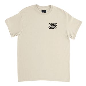 Könnte beinhalten: Cremefarbenes T-Shirt mit einer kleinen schwarzen Grafik einer Katze mit Sonnenbrille in einem Planetenring. Das Shirt hat einen Rundhalsausschnitt und kurze Ärmel. Die Katzen-Grafik befindet sich auf der oberen linken Brust.