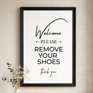 Op de afbeelding: Een ingelijste print met een zwarte rand en een witte achtergrond. De tekst luidt "Welcome, PLEASE REMOVE YOUR SHOES, thank you" in zwarte letters. Een kleine vaas met groen staat in de linker benedenhoek.