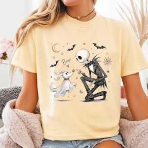 May include: A pale yellow t-shirt featuring a graphic illustration of a skeleton character in a pinstripe suit kneeling to touch noses with a ghost dog. The design includes the text Boop, a crescent moon, bats, stars, and a spider web.