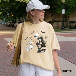 May include: A butter-colored short-sleeve t-shirt featuring a graphic print of a skeleton character kneeling and touching the nose of a ghost dog. The design includes the text "Boop," a crescent moon, bats, stars, and a spider web illustration.