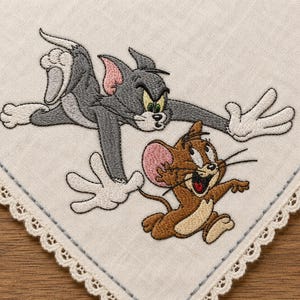 May include: Embroidered handkerchief featuring Tom and Jerry cartoon characters. Tom, a grey cat, is depicted lunging, while Jerry, a brown mouse, is running. The handkerchief has a white background with a decorative lace border.