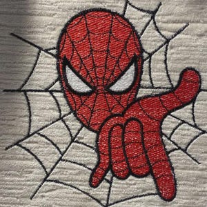 Pode incluir: Um emblema bordado apresentando uma máscara vermelha com detalhes de teia pretos e olhos brancos, posicionado sobre um design de teia de aranha preta costurada em um fundo de tecido branco sujo texturizado. O design mostra um gesto de mão.
