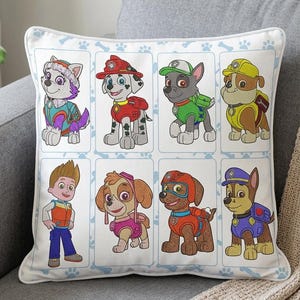 May include: White square throw pillow featuring embroidered images of cartoon dogs and a boy in various outfits. The pillow has a light blue border with paw prints and bone designs. The characters are from the Paw Patrol series.