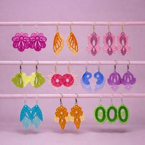 May include: A selection of embroidered earrings in a range of designs and colours, including floral and abstract shapes. The earrings are displayed on pink rods against a pale pink backdrop. Styles include flower, wing, and geometric shapes, in shades of pink, orange, blue, green, and purple.