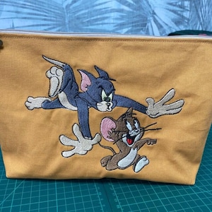 May include: A mustard yellow canvas zippered pouch featuring an embroidered graphic of the cartoon characters Tom and Jerry. The blue cat Tom is reaching out with white-gloved hands toward the brown mouse Jerry, who is running forward.