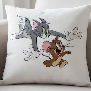 May include: White square cushion with an embroidered design of Tom and Jerry cartoon characters. Tom, a grey cat, is in mid-air, chasing Jerry, a brown mouse. The cushion is on a light-coloured sofa.