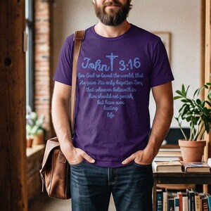 May include: A purple short-sleeved t-shirt featuring the text John 3:16 and a cross graphic in light blue. The shirt displays the full verse text in a script font. The garment is paired with dark blue denim jeans and brown leather shoes.